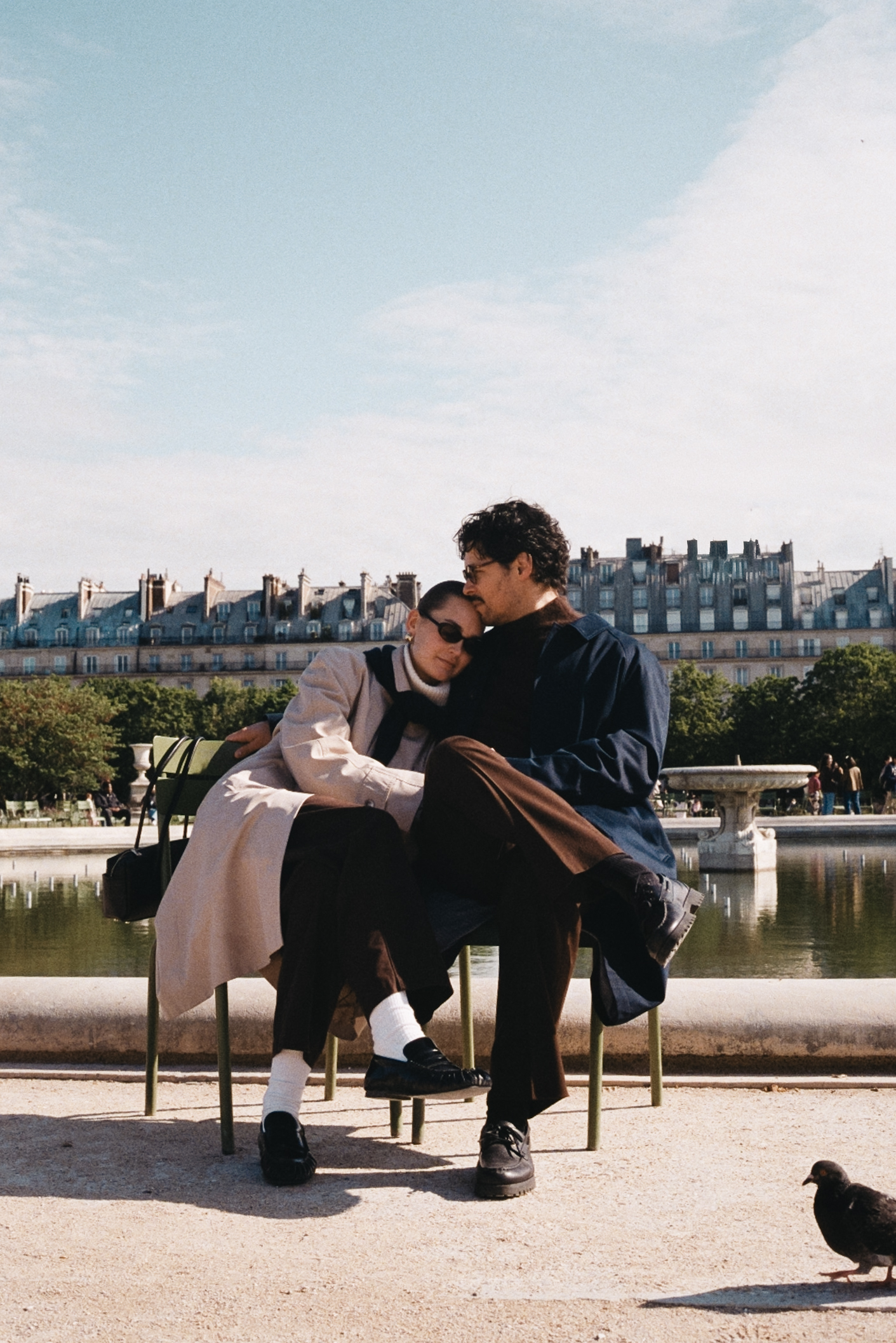 Couple sitting on a bench with pigeons nearby in a Parisian park, wearing coats.