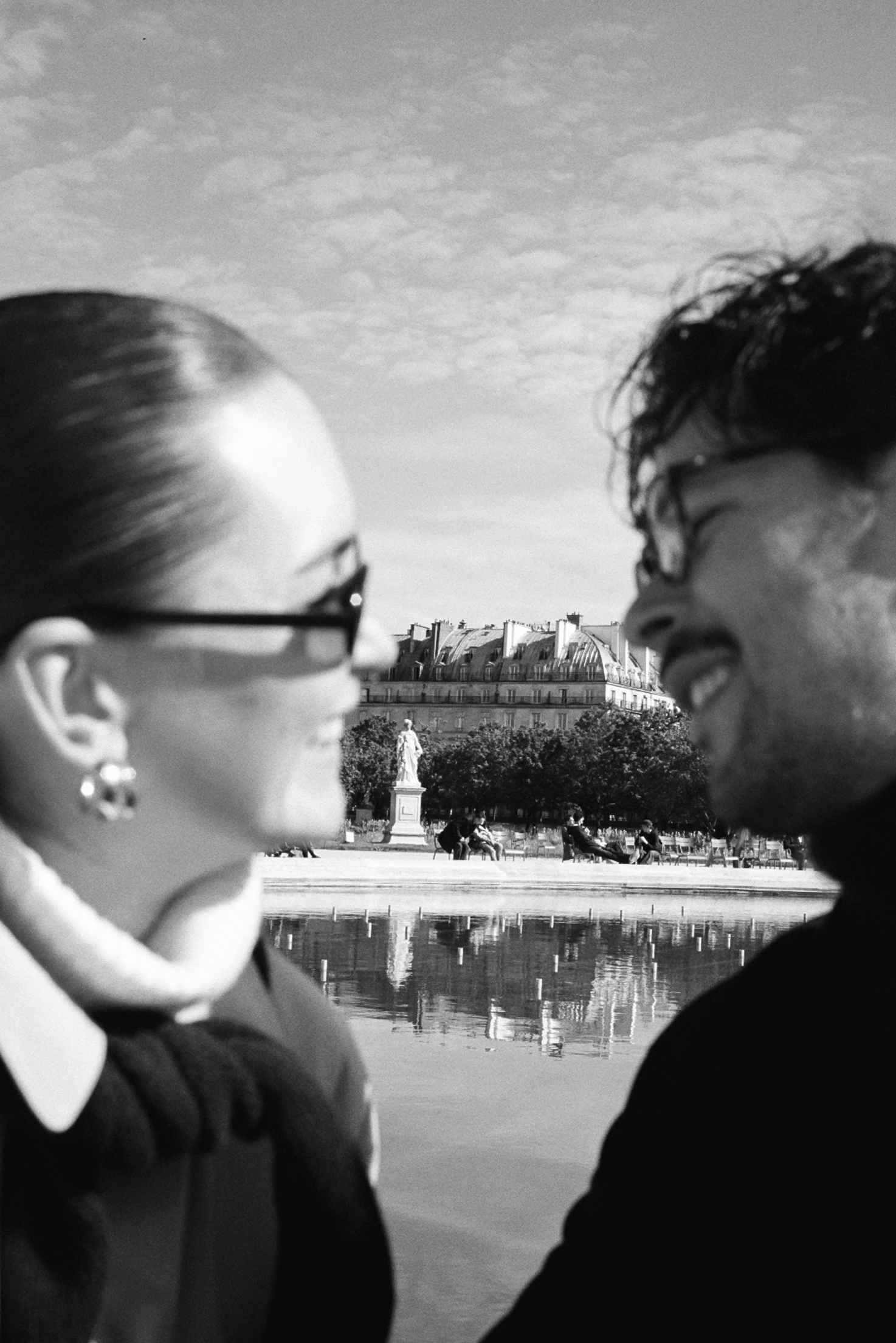Two people in foreground with glasses, close together; cityscape and statue behind.