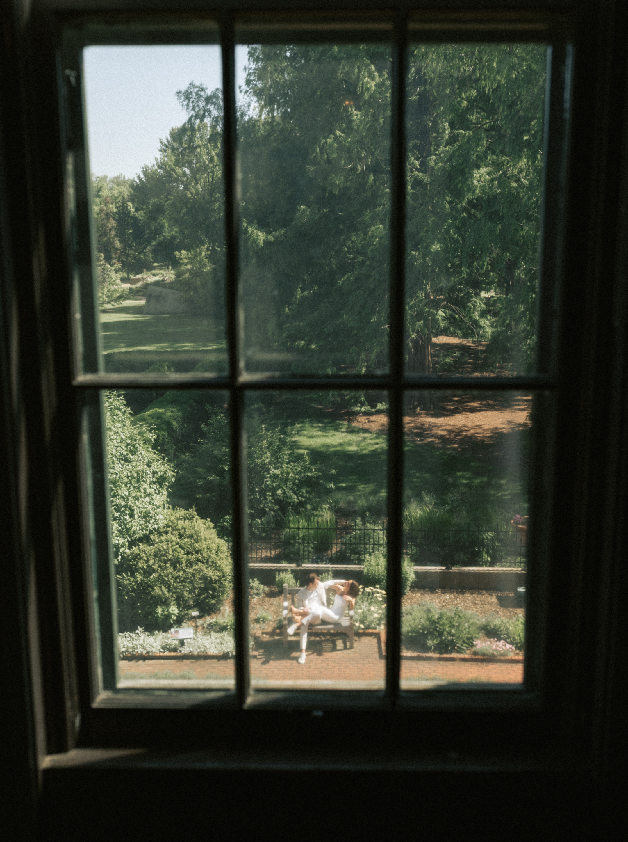 A view through a window looking down at a couple sitting on a bench in a verdant garden.