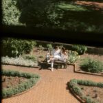 A cinematic view through a window looking down at a couple sitting on a bench in a verdant garden.