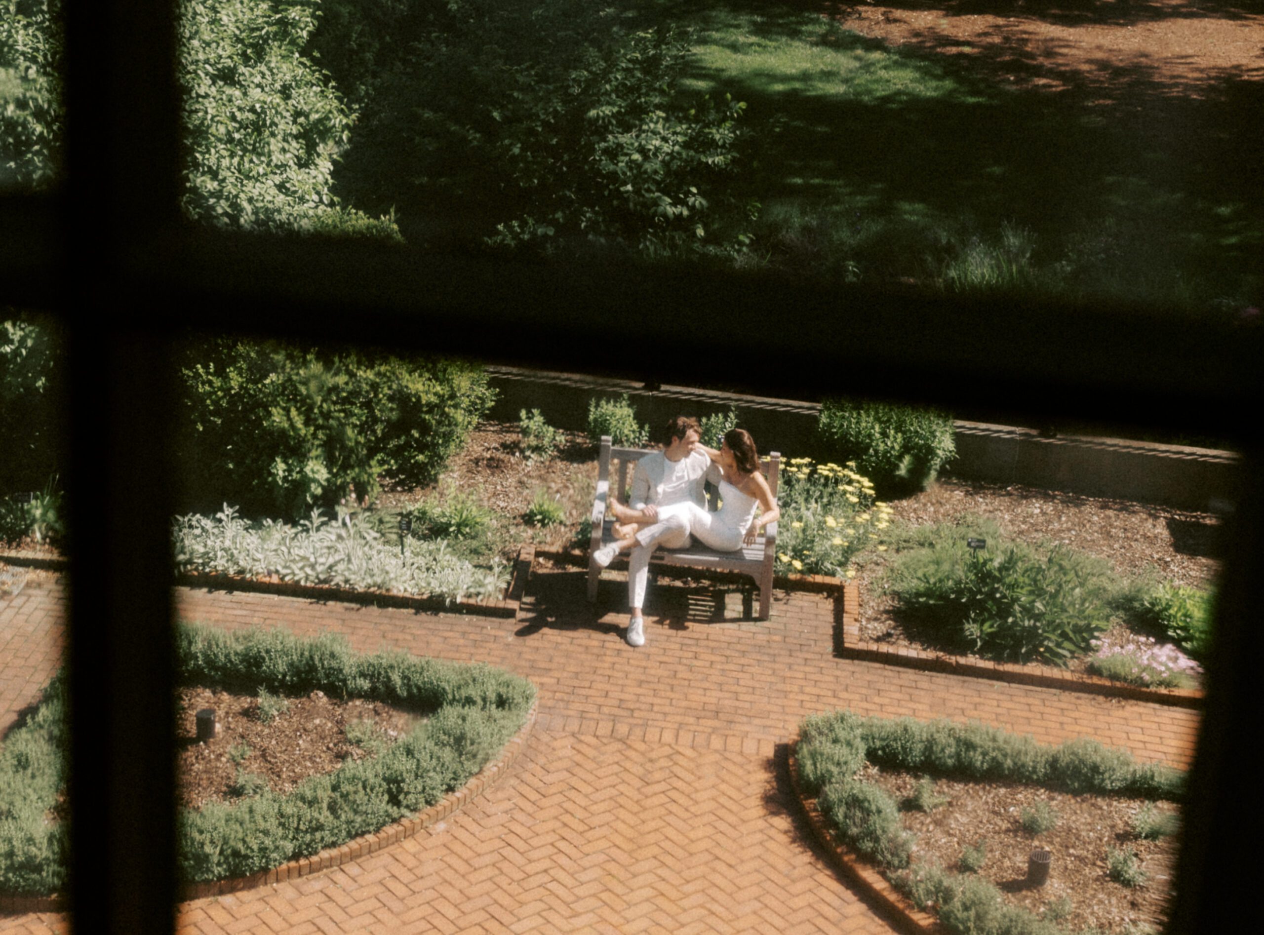 A cinematic view through a window looking down at a couple sitting on a bench in a verdant garden.