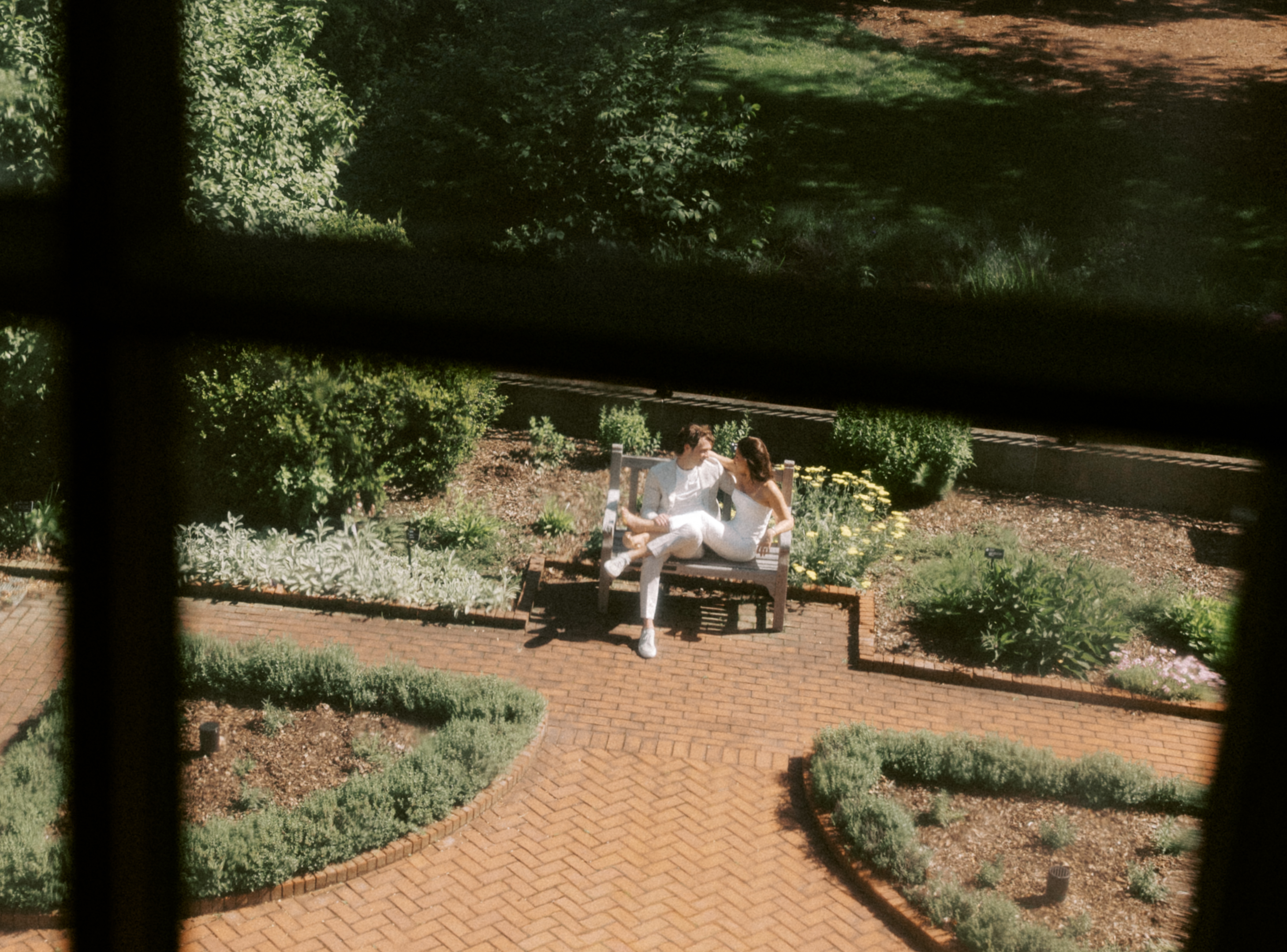 A cinematic view through a window looking down at a couple sitting on a bench in a verdant garden.