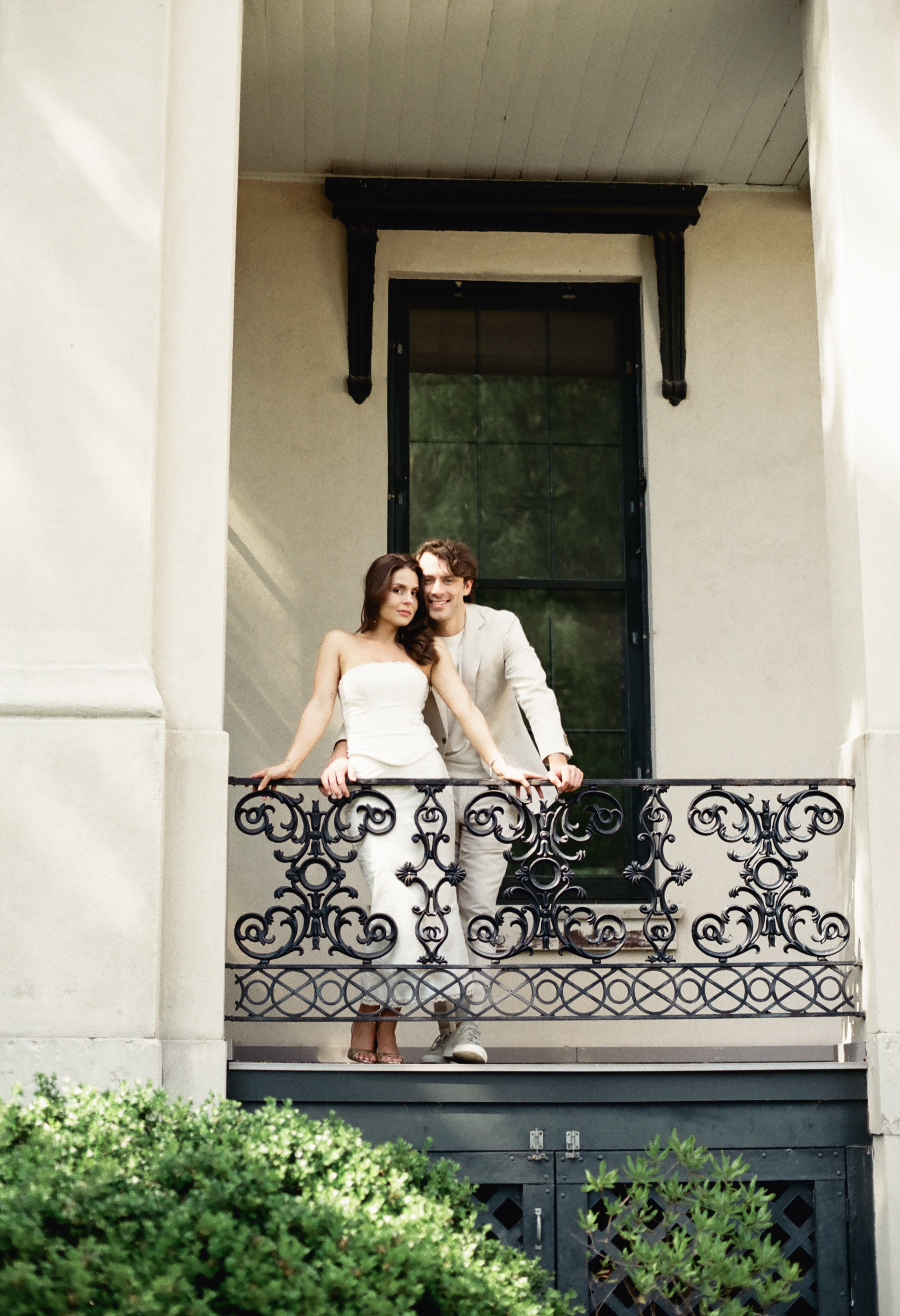 Couple on a decorative balcony in front of a large window, leaning on railing.