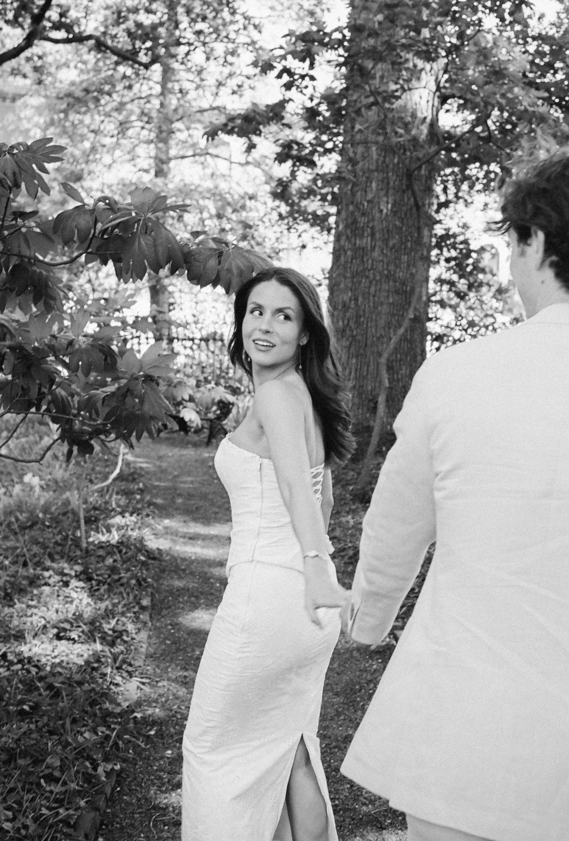35mm film photo of a woman in a white strapless dress stands outdoors, holding hands with a man in a light suit.